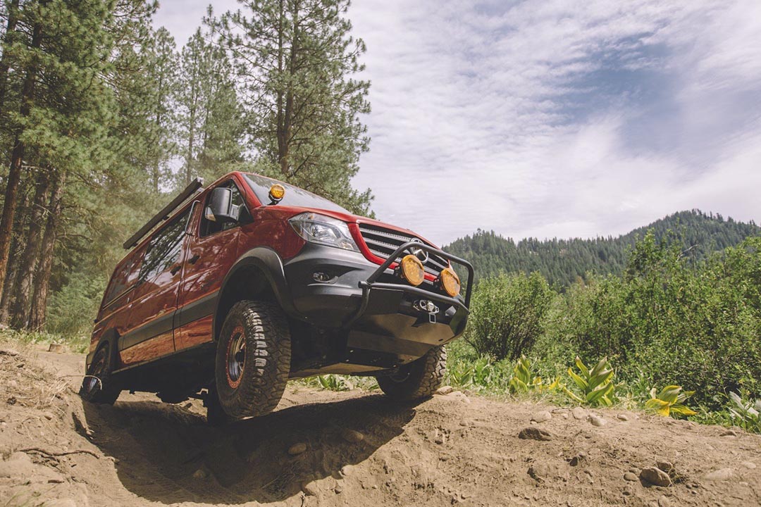 A red truck on a dirt hill, with visible close-ups of its tire and headlight, surrounded by a cloudy sky and trees.