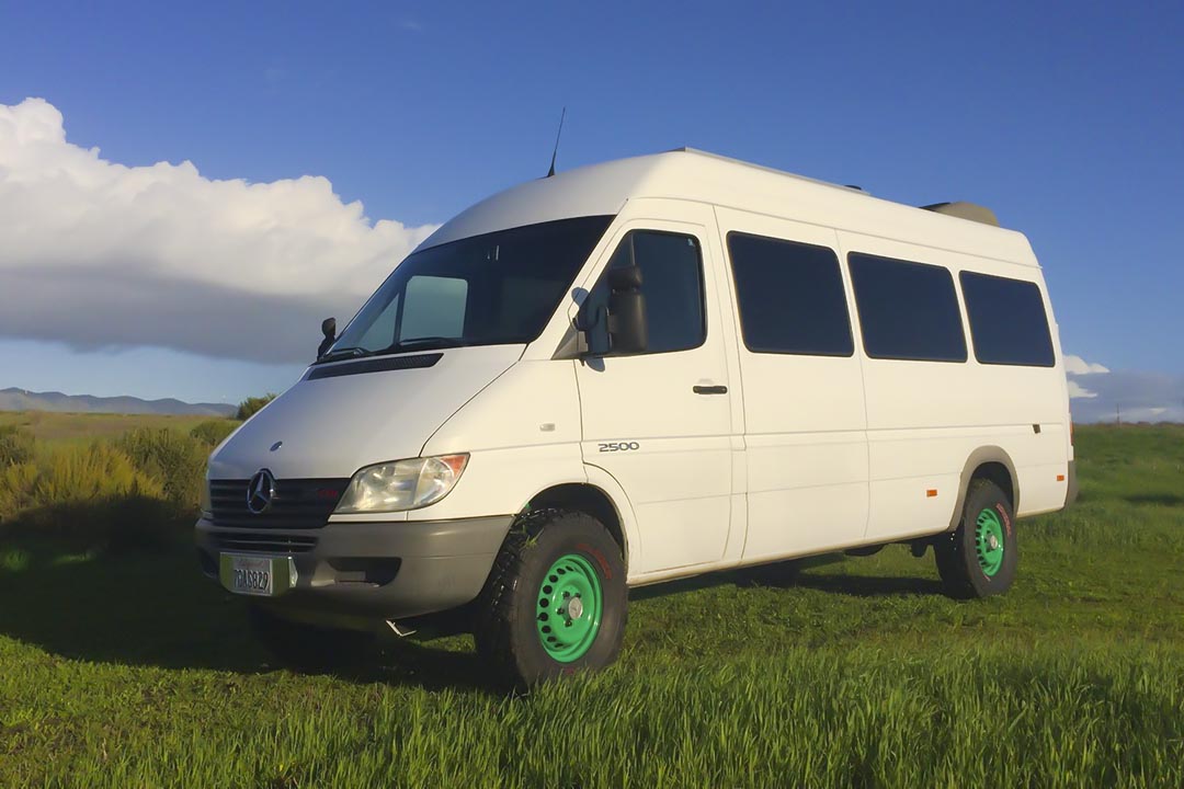 A white van parked on grass with a clear blue sky in the background, showing close-ups of a tire, headlight, and side view mirror.