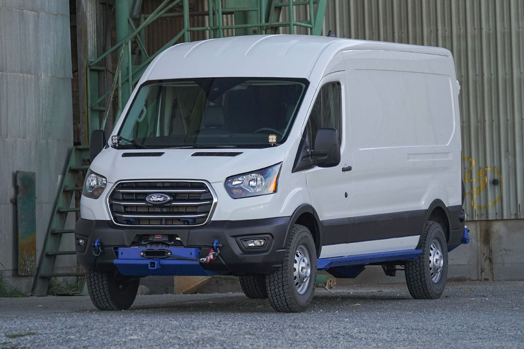 A white van parked in front of a green metal wall, featuring visible headlight, tire, door handle, and car grille.