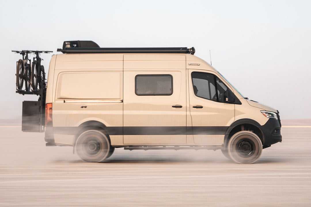 A white van with a black roof drives on a road, showcasing its tires and carrying a bicycle on a rear rack.