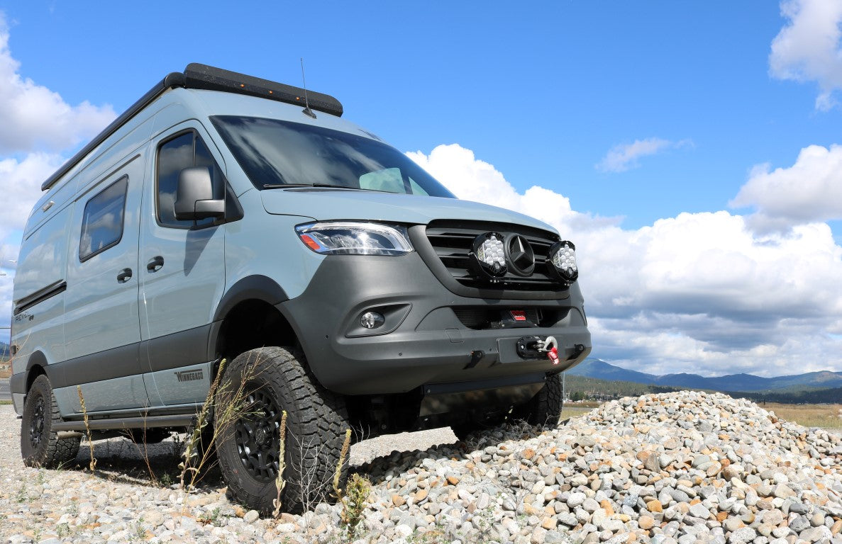 A grey van parked on rocky terrain, showing a close-up of its tire and side view mirror.