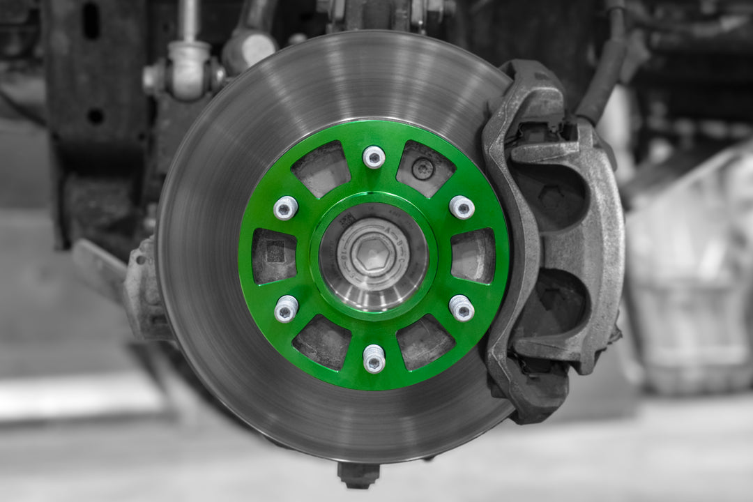 A close-up image of a 14mm wheel spacer, featuring a green anodized aluminum spacer mounted on a vehicle hub, with surrounding components in grayscale for contrast.