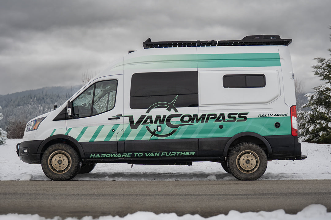 A white Ford Transit camper van with teal and black graphics, a metallic bronze-colored wheel rim with a multi-spoke design and cutouts, featuring the 'Van Compass' brand name and logo embossed on the center cap, parked on a snowy road under an overcast sky. 