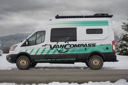 A white Ford Transit camper van with teal and black graphics, a metallic bronze-colored wheel rim with a multi-spoke design and cutouts, featuring the 'Van Compass' brand name and logo embossed on the center cap, parked on a snowy road under an overcast sky. 