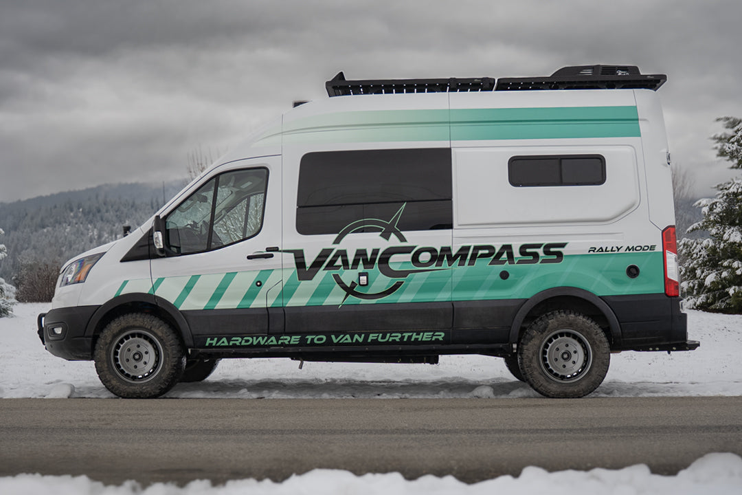 A white Ford Transit camper van with teal and black graphics, with metallic colored wheels that have a bold, multi-spoke design featuring the 'Van Compass' brand name and logo embossed on the center cap, parked on a snowy road under an overcast sky. 
