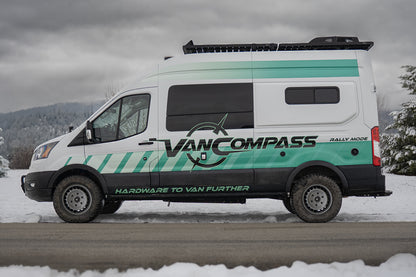 A white Ford Transit camper van with teal and black graphics, with metallic colored wheels that have a bold, multi-spoke design featuring the 'Van Compass' brand name and logo embossed on the center cap, parked on a snowy road under an overcast sky. 