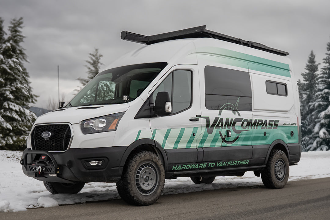 A white Ford Transit camper van with teal and green graphics, featuring 'VAN COMPASS' branding, with metallic colored wheels that have a bold, multi-spoke design featuring the 'Van Compass' brand name and logo embossed on the center cap, parked on a snowy road with pine trees in the background. 