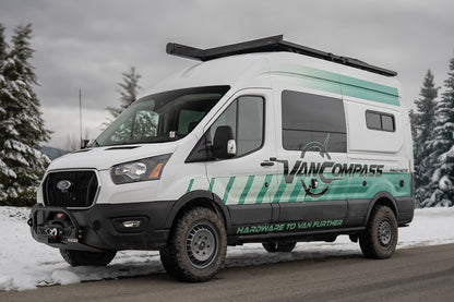 A white Ford Transit camper van with teal and green graphics, featuring 'VAN COMPASS' branding, with metallic colored wheels that have a bold, multi-spoke design featuring the 'Van Compass' brand name and logo embossed on the center cap, parked on a snowy road with pine trees in the background. 