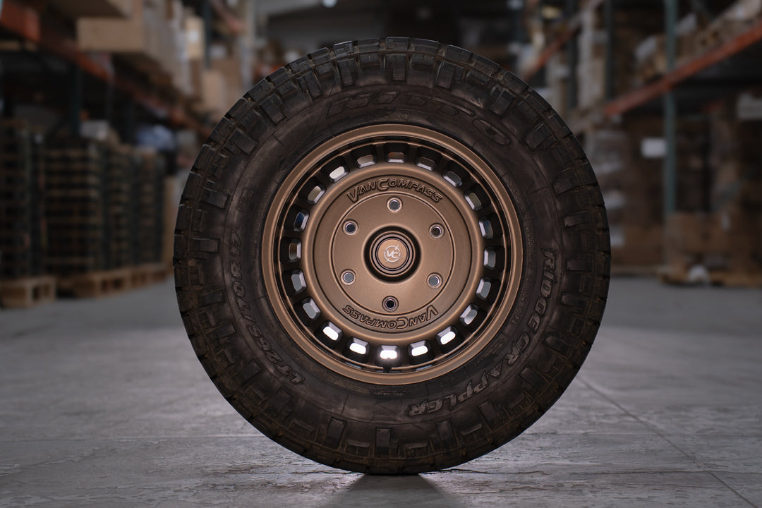 A rugged off-road tire mounted on a bronze-colored multi-spoke wheel, photographed in a warehouse setting with blurred shelving in the background.