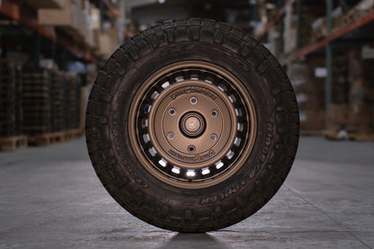 A rugged off-road tire mounted on a bronze-colored multi-spoke wheel, photographed in a warehouse setting with blurred shelving in the background.